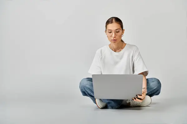 A young woman in a white t-shirt and glasses sitting on the floor, engrossed in her laptop on a grey background. — Stock Photo