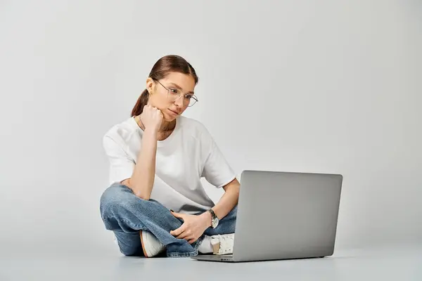 A young woman in a white t-shirt and glasses sits on the floor, focused on her laptop screen against a grey background. — Stock Photo