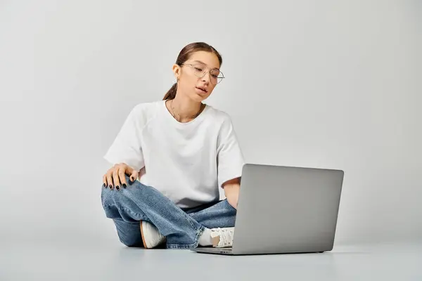 Una mujer joven con una camiseta blanca y gafas se sienta en el suelo, enfocada en la pantalla de su computadora portátil, comprometida con el trabajo o el estudio.. - foto de stock