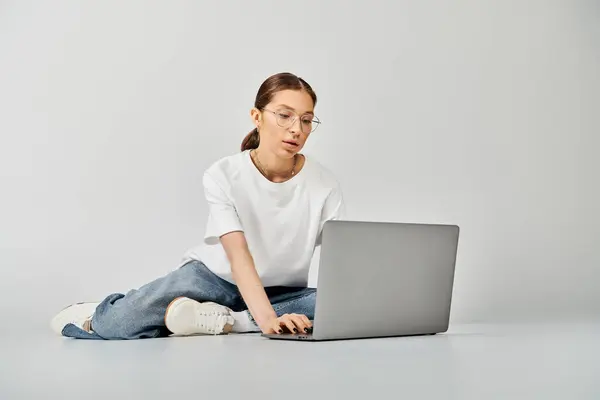 Una giovane donna con una t-shirt bianca e occhiali è seduta sul pavimento, assorta nell'uso del suo computer portatile su uno sfondo grigio. — Foto stock