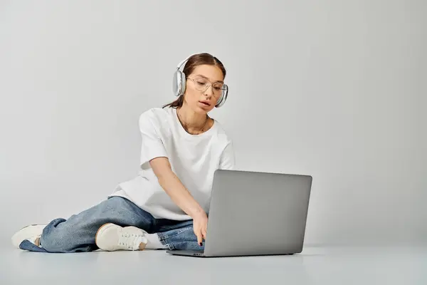 Una giovane donna in t-shirt bianca e occhiali seduta sul pavimento, concentrata mentre lavora su un computer portatile. — Foto stock