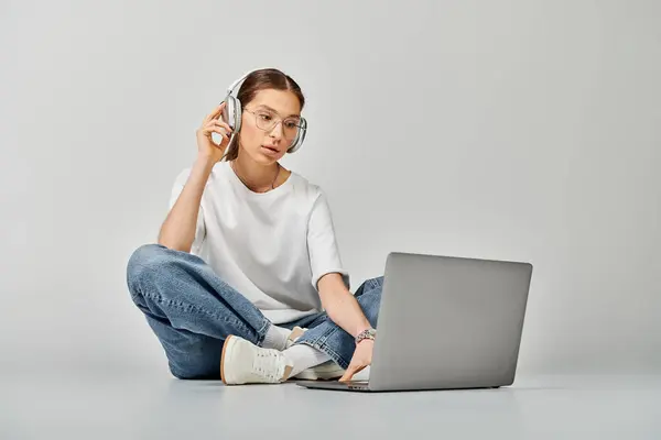 Una joven con una camiseta blanca y gafas se sienta en el suelo con un portátil y auriculares, absortos en su trabajo. — Stock Photo