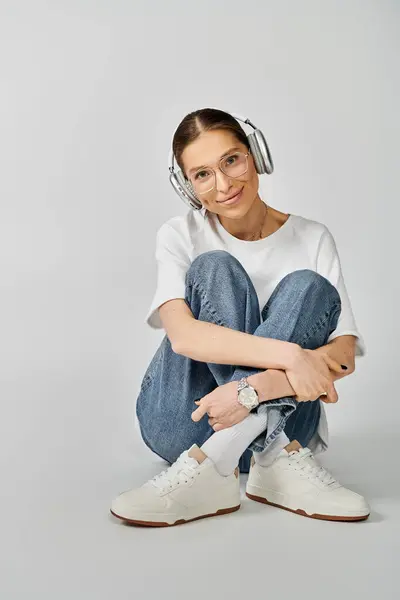 Uma jovem mulher em uma camiseta branca e óculos se senta no chão, imersa em sua música enquanto usa fones de ouvido. — Fotografia de Stock