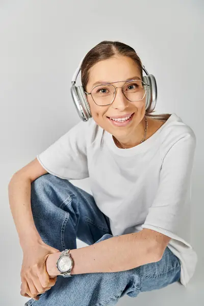 A young woman in a white t-shirt and glasses sits on the floor, immersed in music with headphones on. — Stock Photo
