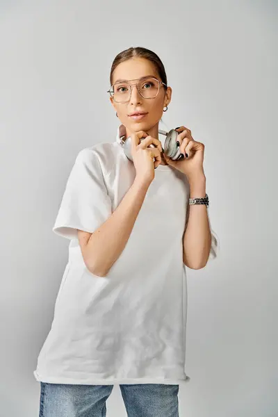 A stylish young woman in a white t-shirt and glasses holding headphones against a grey background. — Stock Photo