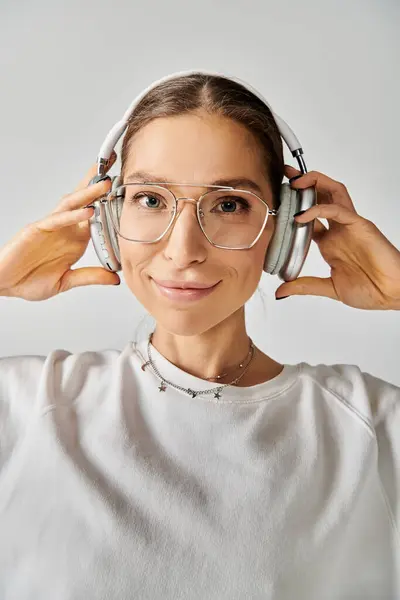Una joven con gafas y auriculares, escuchando atentamente sobre un fondo gris. — Stock Photo
