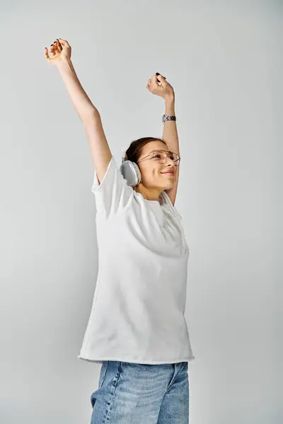 Eine junge Frau in weißem Hemd und Brille hebt jubelnd die Arme vor grauem Hintergrund. — Stockfoto