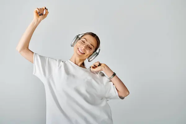 Una giovane donna con le cuffie che indossa una t-shirt bianca, godendo della musica su uno sfondo grigio. — Foto stock