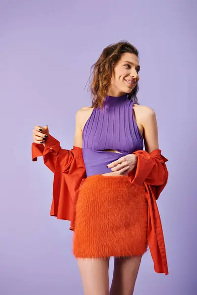 Elegante giovane donna si erge con grazia in top viola e gonna arancione contro uno sfondo viola vibrante. — Foto stock