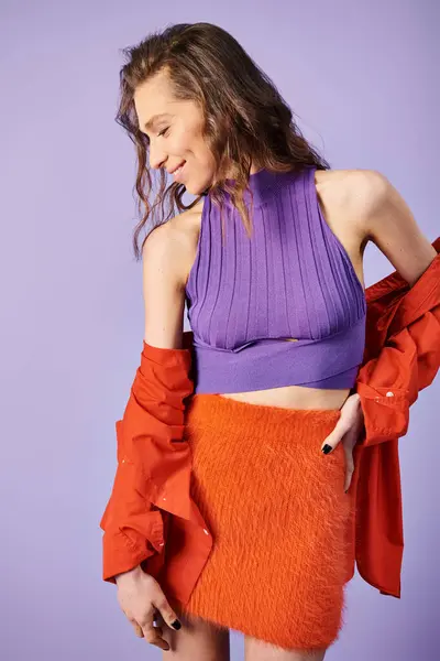 A stylish young woman stands out in a purple top and orange skirt against a vibrant purple background. — Stock Photo