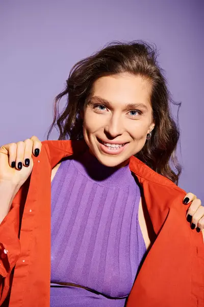 A fashionable young woman wearing a purple top and vibrant orange jacket standing against a purple background. — Stock Photo