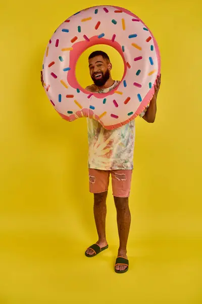 Ein Mann hält sich spielerisch einen riesigen Donut vor das Gesicht, der ihn komplett bedeckt. Die bunten Streusel stehen im Kontrast zu seinem Ausdruck der Freude. — Stockfoto
