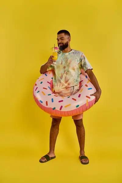 Um homem vestindo uma camisa de gravata vibrante está alegremente segurando uma bebida refrescante em uma mão e um divertido donut flutuar na outra.. — Fotografia de Stock