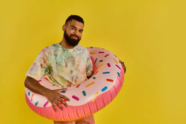 Un uomo che tiene gioiosamente una ciambella enorme davanti a uno sfondo giallo vibrante, mostrando il trattamento zuccherino. — Foto stock