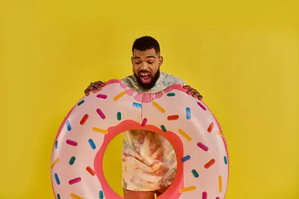 Мужчина с улыбкой на лице, держащий массивный пончик, покрытый красочными брызгами, демонстрирующий чувство радости и снисходительности в сюрреалистический момент. — стоковое фото