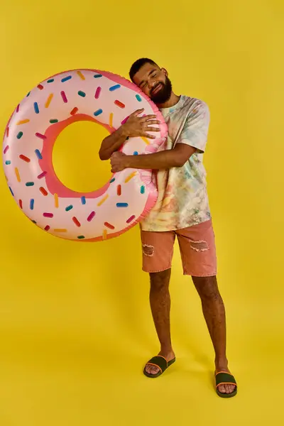 Мужчина радостно держит гигантский пончик перед ярким желтым фоном, демонстрируя свою любовь к сладкому угощению. — стоковое фото