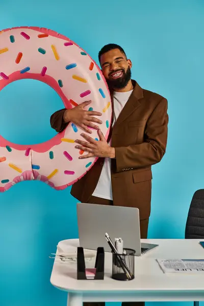 Um homem está segurando um donut enorme na frente de um laptop, aparentemente interagindo com a tela. A justaposição do doce deleite e da tecnologia cria uma cena caprichosa e surreal. — Fotografia de Stock