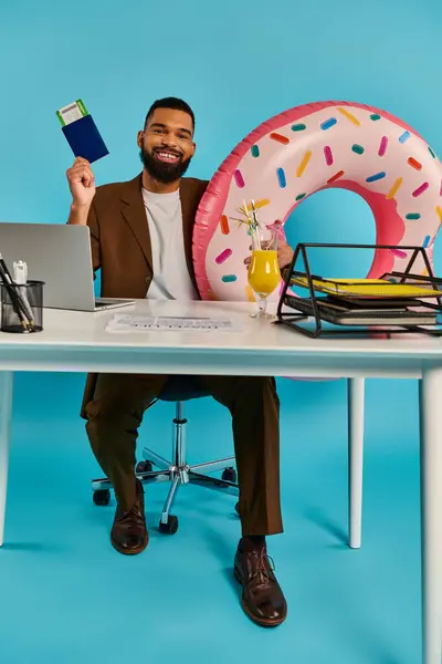 Un hombre sentado en un escritorio de madera, enfocado en su computadora portátil mientras disfruta de un delicioso donut con chispas. - foto de stock