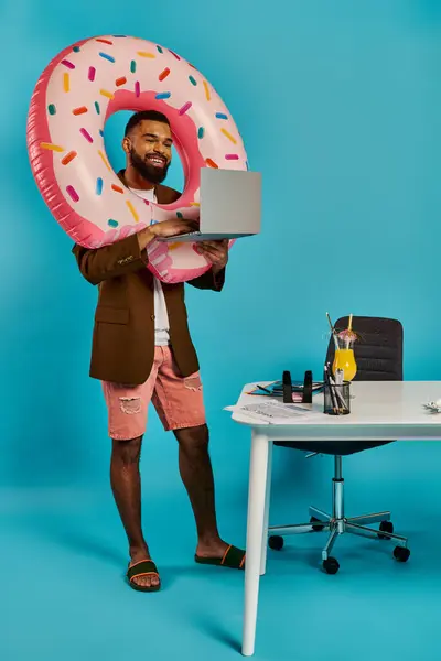 Un uomo tiene un computer portatile in una mano e una ciambella gigante nell'altra, mostrando un equilibrio di lavoro e giocare in un ambiente stravagante. — Foto stock