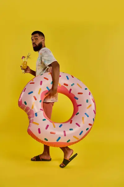 Un hombre sostiene alegremente un donut gigantesco en una mano y un vaso de cerveza en la otra, mostrando una combinación única y deliciosa de golosinas. - foto de stock