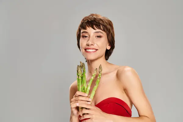 A captivating young woman in a radiant red dress holds a bunch of fresh asparagus. — Stock Photo