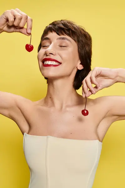 Young woman in a white dress gracefully holds a single cherry against a vibrant backdrop. — Stock Photo