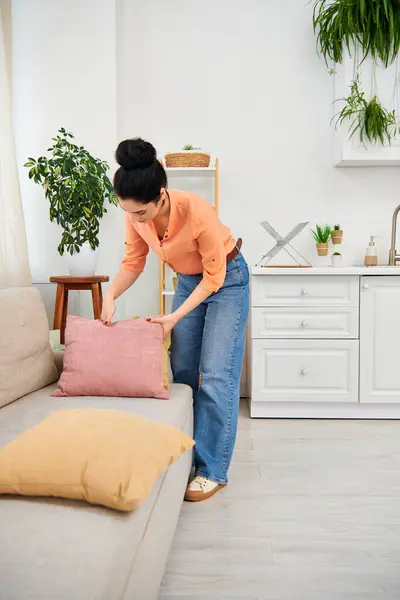 Una donna elegante in abbigliamento casual posiziona con attenzione un cuscino decorativo su un divano moderno, aggiungendo un tocco accogliente al suo spazio vitale.. — Foto stock
