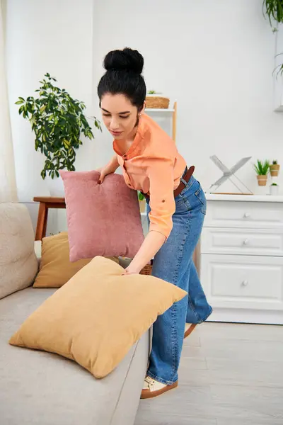 A stylish woman in casual attire arranges a pillow on her couch, creating a cozy and inviting atmosphere in her home. — Stock Photo