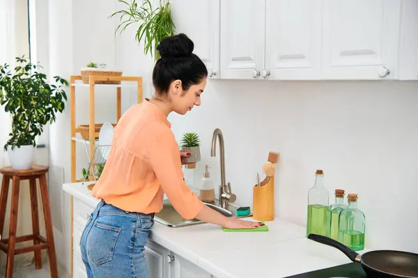 Una donna in abbigliamento casual si trova in un lavandino da cucina, con una padella sul bancone. Sembra concentrata e serena mentre svolge le sue faccende domestiche. — Foto stock