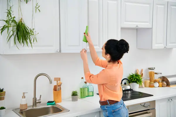 A stylish woman in casual attire methodically scrubs the kitchen sink with a vibrant green rag, bringing radiant cleanliness. — Stock Photo