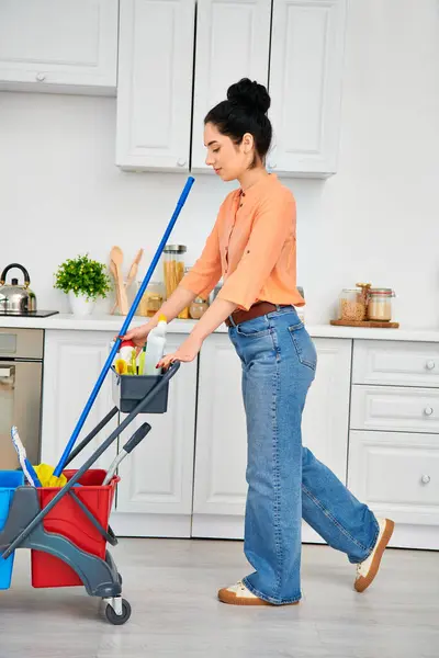 A stylish woman in casual attire pushes a stroller through a cluttered kitchen while multitasking at home. — Stock Photo
