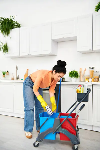 Una mujer elegante con atuendo casual limpia meticulosamente el piso de su cocina con un cubo, absorta en perfeccionar cada esquina. - foto de stock