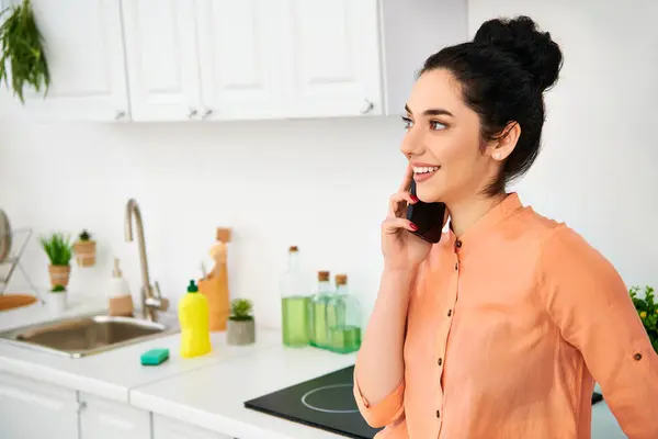 A stylish woman in casual attire stands in a kitchen, chatting on a cell phone while working. — Stock Photo