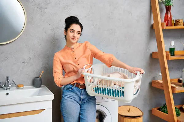 A stylish woman in casual attire holding a laundry basket standing next to a washer, preparing to do laundry. — Stock Photo