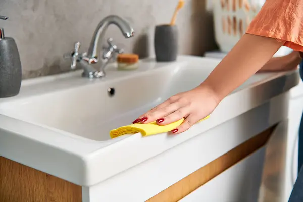 A stylish woman in casual attire diligently scrubs a sink with a bright yellow sponge to remove dirt and achieve a spotless shine. — Stock Photo