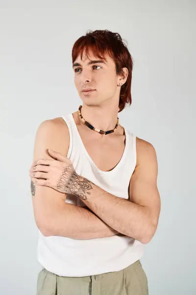 Модный молодой человек с яркими рыжими волосами наносит удар по позе в белом топе на фоне серой студии. — стоковое фото
