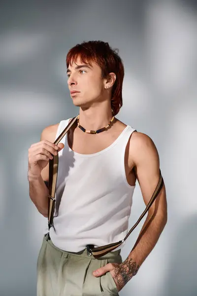Ein stilvoller junger Mann in weißem Tanktop steht vor grauem Hintergrund und hält einen Hosenträger in der Hand. — Stockfoto