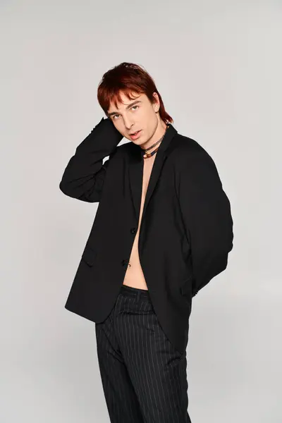 Un joven elegante con el pelo rojo golpea una pose en una chaqueta negra contra un fondo gris en un ambiente de estudio. - foto de stock