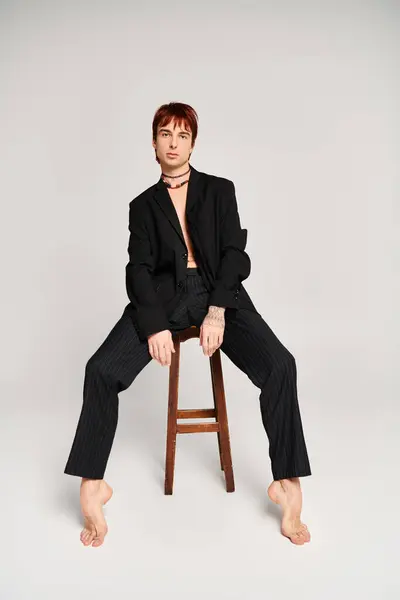 A stylish young man gracefully poses while sitting on top of a wooden stool in a studio with a grey background. — Stock Photo