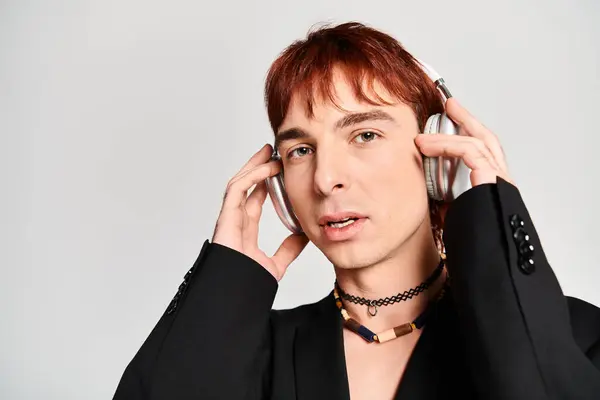 A stylish young man with red hair wearing a black suit and holding a pair of headphones in a studio with a grey background. — Stock Photo