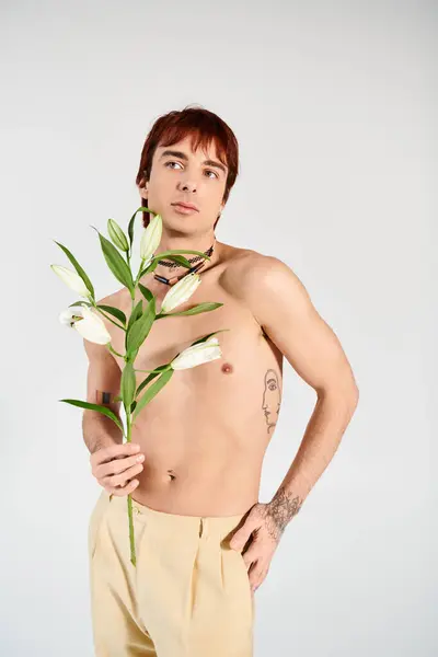 Ein Mann ohne Hemd hält friedlich eine Blume in einem Atelier vor grauem Hintergrund. — Stockfoto
