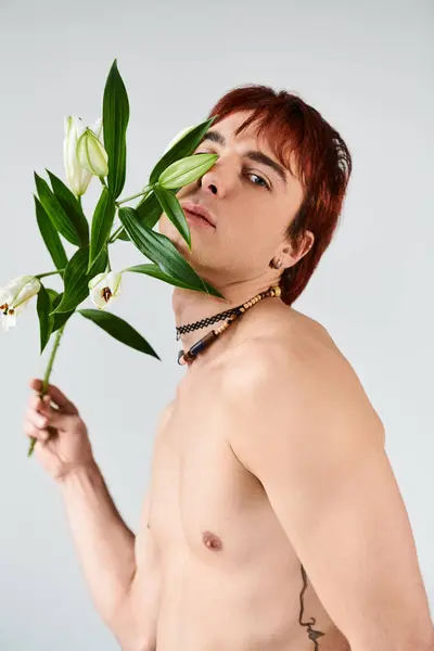 Um homem jovem e sem camisa segura pacificamente uma flor delicada em um estúdio com um fundo cinza. — Fotografia de Stock