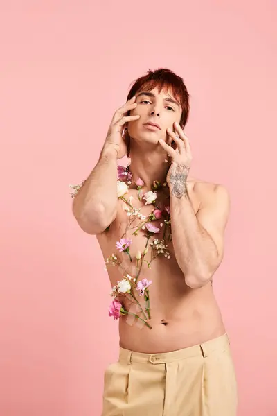 Um jovem sem camisa delicadamente exibe flores em seu peito, marcando uma pose cativante em um estúdio contra um pano de fundo rosa.. — Fotografia de Stock