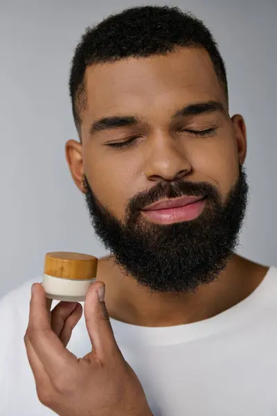 Un bel homme avec une barbe tenant un pot de crème pour sa routine de soins de la peau. — Photo de stock