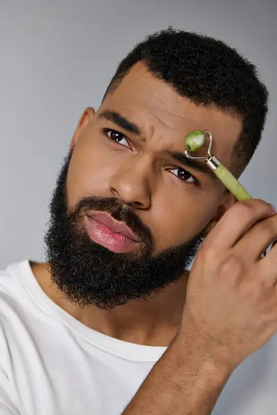 African american bearded man using face roller a for skincare routine. — Stock Photo