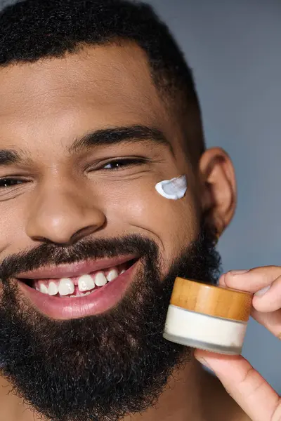 Bearded man joyfully holds jar of cream during skincare routine. — Stock Photo