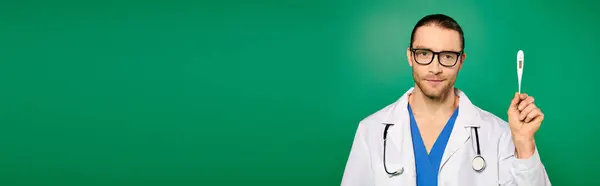Médico bonito em casaco branco segurando um termômetro contra um pano de fundo verde. — Fotografia de Stock