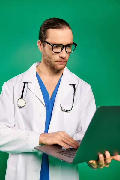 Handsome doctor in white lab coat working on laptop against green backdrop. — Stock Photo