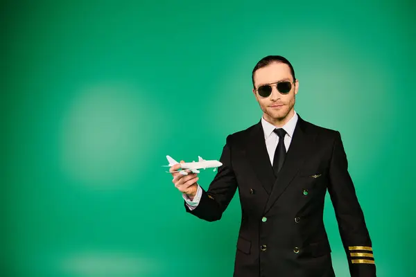 Handsome man in black suit and sunglasses holding model airplane over green backdrop. - foto de stock
