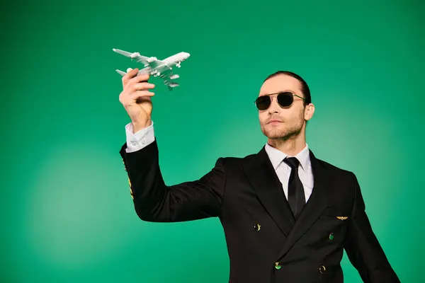 Handsome pilot in black suit holding model airplane against green backdrop. - foto de stock